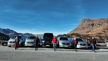 Eine Gruppe von Menschen steht vor mehreren Autos in einer bergigen Landschaft. Der Himmel ist blau und die Umgebung ist sonnig.