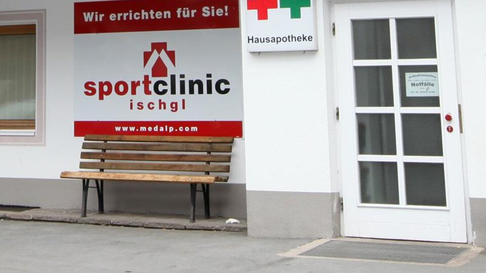 Eine Sportklinik mit einem roten Schild und einer Bank im Freien. Nebenan befindet sich ein Apothekenzeichen.