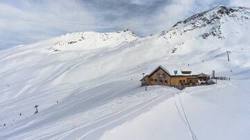 A mountain cabin in the snow, surrounded by snow-covered mountains. The landscape appears calm and wintry.