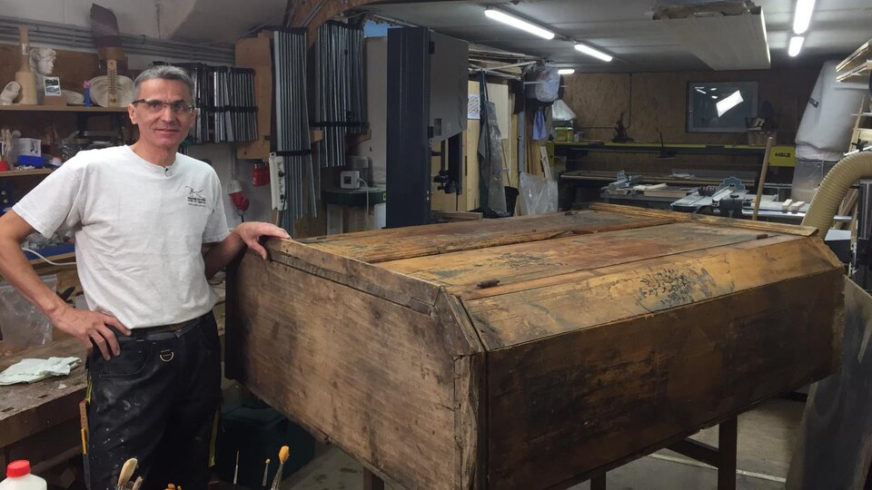 Ein Handwerker steht in seiner Werkstatt neben einer großen, alten Holzkiste. Im Hintergrund sind verschiedene Werkzeuge und Maschinen zu sehen.