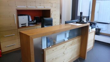 Ein moderner Empfangsbereich mit Holzverkleidung und einem Schreibtisch. Im Hintergrund sind Bürogeräte und ein großer Fensterbereich zu sehen.