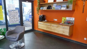 Ein modernen Empfangsbereich mit orangefarbener Wand, einem Holzregal und einem bequemen grauen Sessel. Auf dem Regal befinden sich Informationsmaterialien und eine Kaffeemaschine.