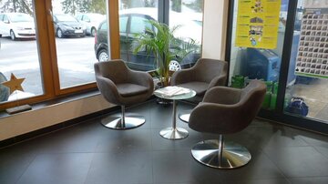 Ein modernes Wartezimmer mit drei grauen Sesseln und einem kleinen Tisch. Durch große Fenster sind Autos und eine grüne Pflanze sichtbar.