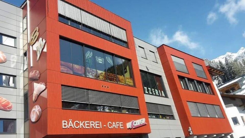 Ein modernes Gebäude mit auffälliger roter Fassade. Im Erdgeschoss befinden sich eine Bäckerei und ein Café.