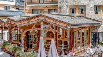 A cozy restaurant with wooden decor and patio furniture. Surrounded by flowers and a picturesque mountain panorama.
