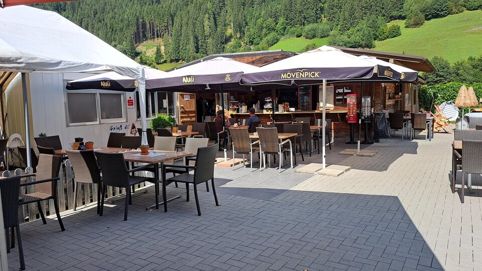 Ein gemütlicher Außenbereich eines Restaurants mit Tischen und Stühlen. Im Hintergrund befindet sich das Restaurant mit Schirmen und einer grünen Landschaft.
