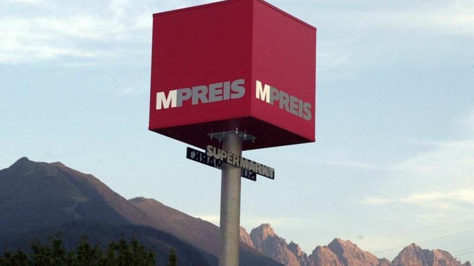 Ein rotes Werbeschild mit der Aufschrift "MPREIS" steht vor einem Gebirgshorizont. Der Schild zeigt auf eine Straße mit dem Zusatz "Supermarkt".