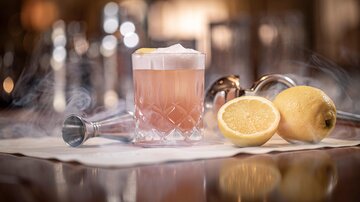 An elegant cocktail in a glass chalice, surrounded by steaming ice. Next to the drink, fresh lemon halves rest on a white table.