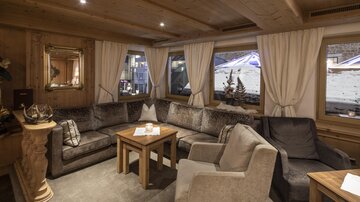 A cozy living area with a large U-shaped sofa and a wooden table. Through the windows, a winter landscape can be seen.