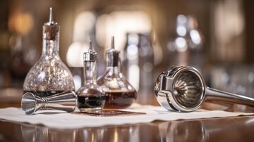 An elegant bar with various glasses and utensils for cocktail preparation. In the foreground, a milk frother and a measuring cup can be seen.