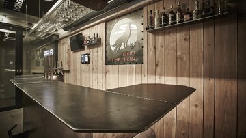 Eine stilvolle Bar mit einem hölzernen Design und einer metallenen Theke. Regale mit Spirituosen und eine Wanddekoration verleihen dem Raum eine einladende Atmosphäre.