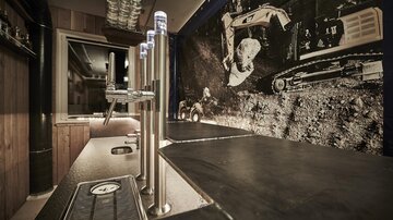 Eine moderne Bar mit einem langen Tresen und Zapfhähnen. Im Hintergrund ist ein Fenster mit einem Bild von Baumaschinen und Bergbau zu sehen.