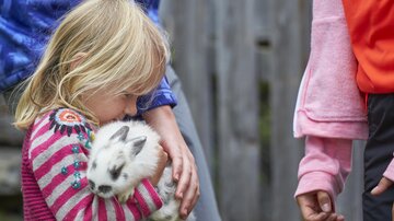 Das Bild zeigt ein blondes Kind, das ein weiß-grau geflecktes Kaninchen eng und vorsichtig im Arm hält. Das Kind trägt einen bunt gemusterten Pullover und beugt den Kopf sanft zum Tier hinunter, wodurch der Moment sehr ruhig und liebevoll wirkt.

Rechts im Bild ist ein weiteres Kind nur teilweise zu sehen, im Hintergrund stehen Erwachsene oder ältere Personen unscharf daneben. Der Fokus liegt klar auf der Nähe zwischen Kind und Kaninchen. Die Szene vermittelt Geborgenheit, Fürsorge und kindliche Zuneigung zu Tieren.