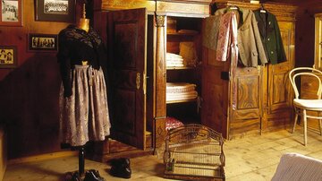Ein gemütlicher Raum mit einem alten Kleiderschrank, einer Puppe in traditioneller Tracht und verschiedenen Kleidungsstücken, die daran hängen. Der Holzboden und die Wände verleihen dem Raum eine warme Atmosphäre.