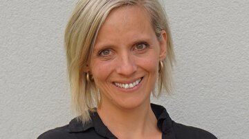 A smiling woman with blonde hair stands in front of a bright wall. She is wearing a black top and looks friendly.