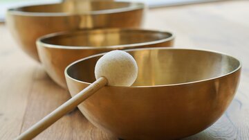 Three metal singing bowls are placed on a wooden floor. A mallet with a soft head lies next to one of the bowls.