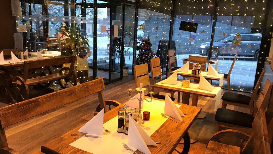Ein gemütliches Restaurant mit Holztischen und -stühlen. Durch große Fenster sieht man weihnachtliche Dekorationen und stimmungsvolles Licht.