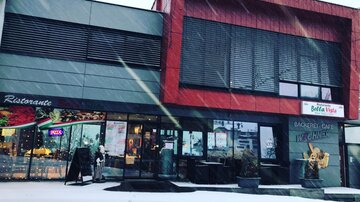 Ein modernes Restaurantgebäude mit roter und grauer Fassade im Schnee. Vor dem Eingang sind einige Stühle und Tische sowie Dekorationen sichtbar.