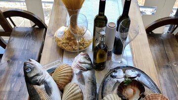 Ein Tisch mit frischen Fischen, Muscheln und Scampi, umgeben von Spaghetti und Olivenöl. Die Anordnung ist appetitlich und zeigt verschiedene Meeresfrüchte.