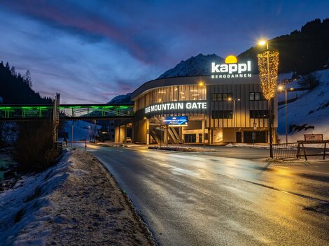 A modern building with the inscription "Kappl Bergbahnen" stands in the foreground. The scene shows a wintry environment at dusk.
