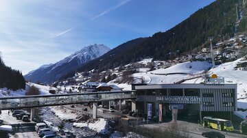 Ein schneebedecktes Tal mit hohen Bergen im Hintergrund. Im Vordergrund sieht man ein modernes Gebäude mit einer Brücke und Autosparkplätzen.