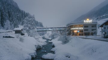 Ein verschneites Winterlandschaft mit einem Fluss und Bäumen. Im Hintergrund steht ein modernes Gebäude mit einem leuchtenden Schild.