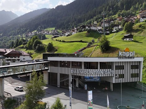A modern building with the inscription "Kappl" in a mountainous landscape. In the background, green hills and small houses can be seen.