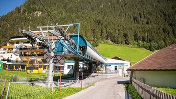 Eine Bergbahnstation umgeben von grünen Wiesen und Wäldern. Im Hintergrund sind alpinen Häuser zu sehen.