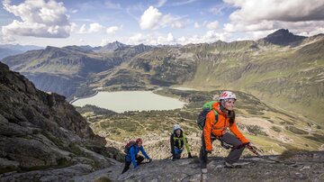 Eine Gruppe von Bergsteigern erklimmt einen Felsen in den Alpen. Im Hintergrund ist ein klarer See und beeindruckende Berglandschaften zu sehen.