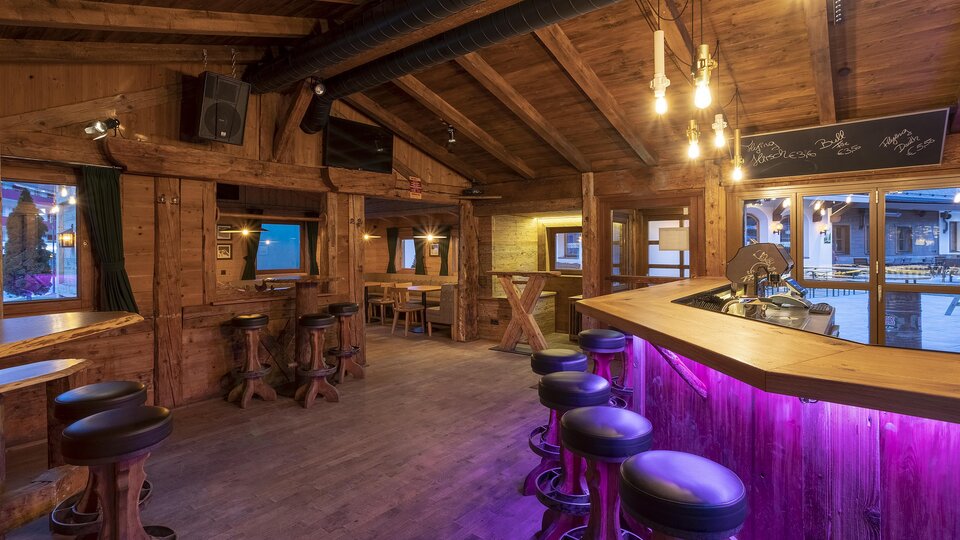 A cozy bar area with wooden interior and soft lighting. The bar has a modern, purple-lit surface and some seating.