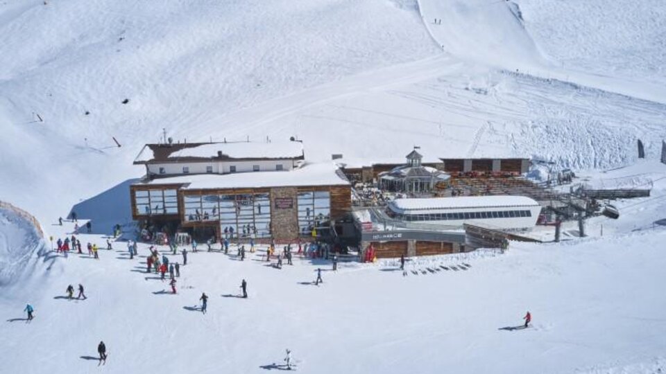 Ein Skigebiet mit viel Schnee und einem großen Holzgebäude. Zahlreiche Skifahrer und Snowboarder genießen die winterliche Landschaft.