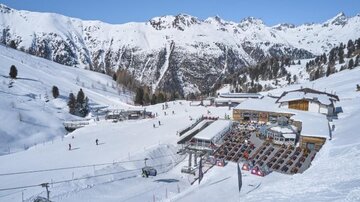 Eine wunderschöne Winterlandschaft mit schneebedeckten Bergen. Im Vordergrund befinden sich eine Skihütte und Liftanlagen, umgeben von Skifahrern.