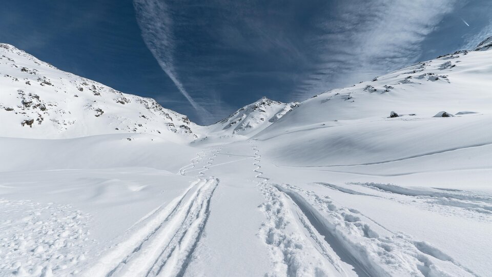 Eine verschneite Landschaft mit Spuren im Schnee und hohen Bergen. Der Himmel ist blau mit einigen Wolken.
