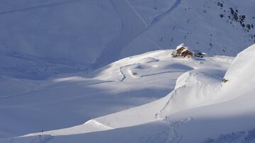 Eine schneebedeckte Landschaft mit sanften Hügeln und einer kleinen Hütte. Klare blaue Himmel und verschneite Berge schaffen eine ruhige Atmosphäre.