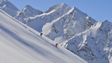 Zwei Skifahrer fahren einen schneebedeckten Hang hinunter. Im Hintergrund sind beeindruckende, schneebedeckte Berge zu sehen.