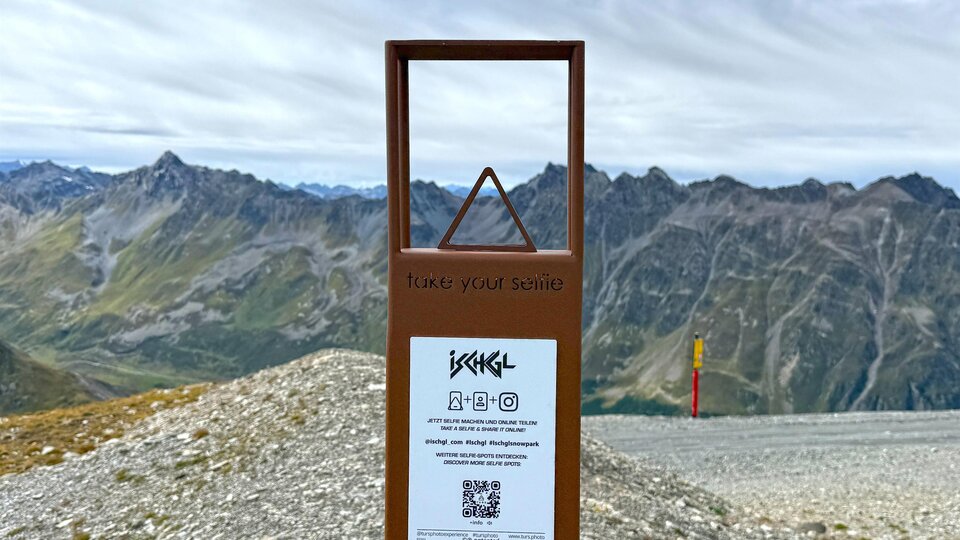 An information sign on a mountain path with a view of the surrounding mountains. The sky is cloudy and the landscape is rocky.