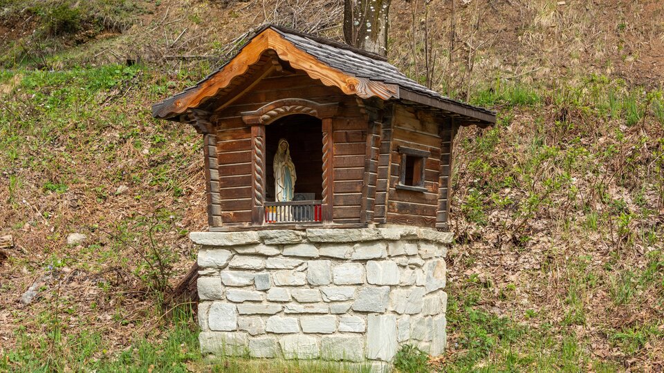 Eine kleine Holzkapelle steht auf einem Steinsockel in der Natur. Neben der Kapelle sind grüne Wiesen und Laub zu sehen. | © Bernhard Gruber – Photography