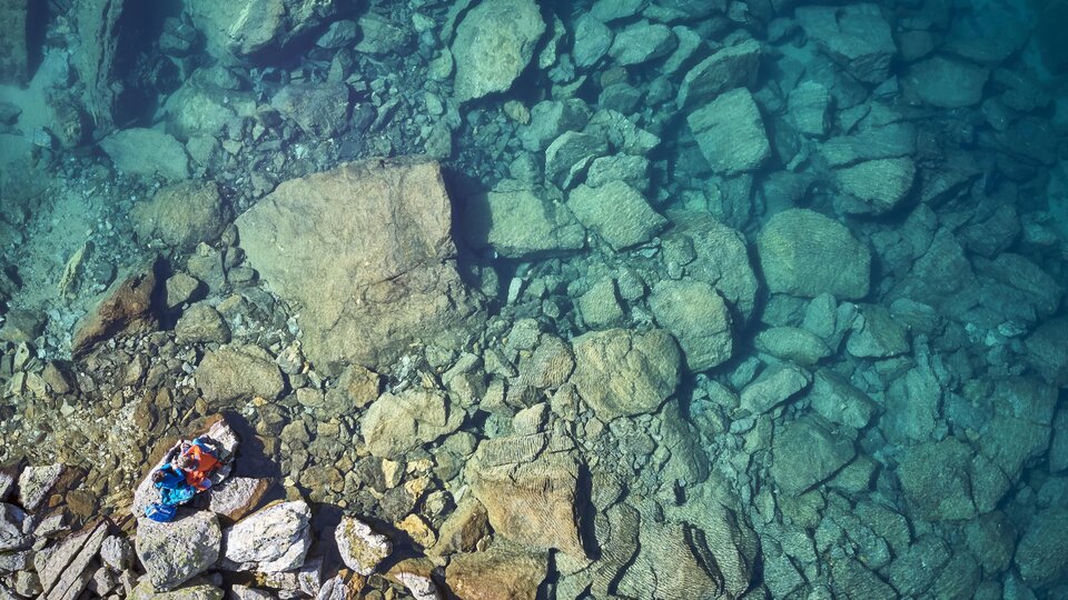 Ein klarer, blauer See mit vielen großen Steinen am Grund. Eine Person liegt entspannt am Ufer.