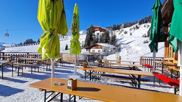 Eine gemütliche Terrasse mit Holztischen und bunten Sonnenschirmen im Schnee. Im Hintergrund sind verschneite Berge und eine Seilbahn zu sehen.