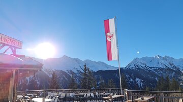 Ein schöner Ausblick auf schneebedeckte Berge unter einem klaren blauen Himmel. Im Vordergrund stehen Tische und eine Fahne weht im Wind.