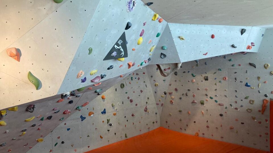 A climbing wall in an indoor space with many colorful holds. The floor is covered with an orange mat.