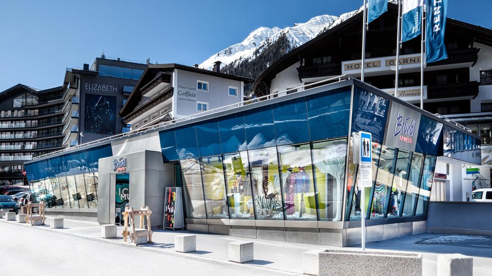 Ein modernes Gebäude mit großen Glasfenstern in einer Alpenlandschaft. Im Hintergrund sind schneebedeckte Berge und typische alpine Architektur zu sehen.