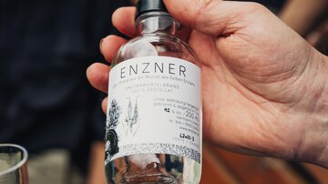 Eine handgehaltene Flasche mit dem Etikett „ENZNER“ steht im Vordergrund. Im Hintergrund sind verschwommen Menschen und eine Holzbank zu sehen.