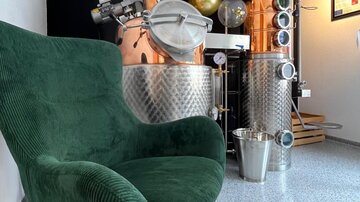 Ein gemütlicher grüner Sessel steht vor einer glänzenden Kupfer- und Edelstahl-Destillationsanlage. Der Raum hat ein modernes Ambiente mit einer klaren, minimalistischen Einrichtung.
