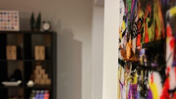 Ein bunter Wandbehang mit einem künstlerischen Motiv hängt an der Wand. Im Hintergrund sind Regale mit verschiedenen Gegenständen zu sehen.