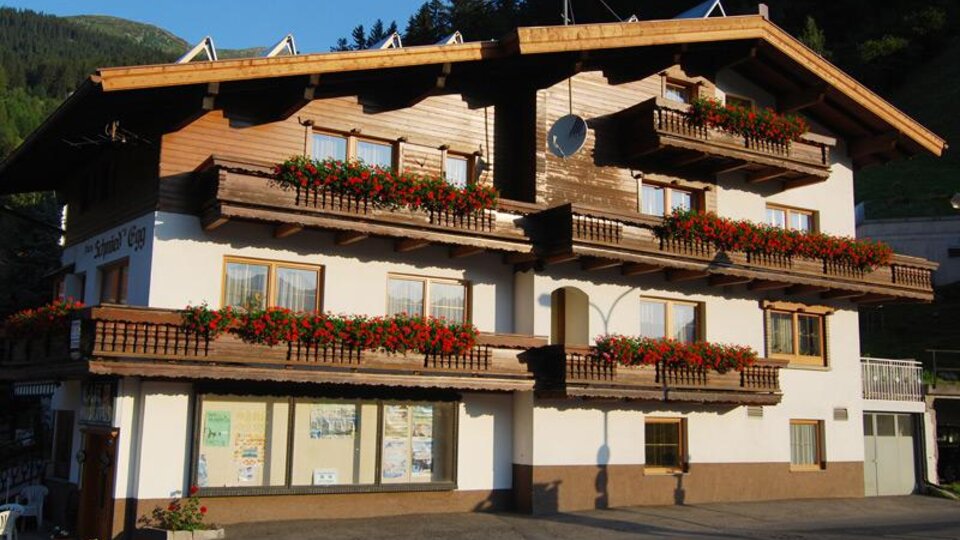 A traditional wooden building with many balconies adorned with red flowers. In the background, green mountains can be seen.