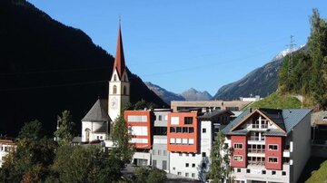 A picturesque mountain village with a church and modern buildings. The surroundings are characterized by green trees and high mountains.