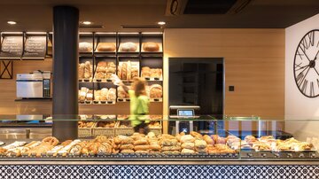 Eine moderne Bäckerei mit einer Auswahl an frischem Brot und Gebäck. Im Hintergrund ist eine Mitarbeiterin zu sehen, die arbeitet.