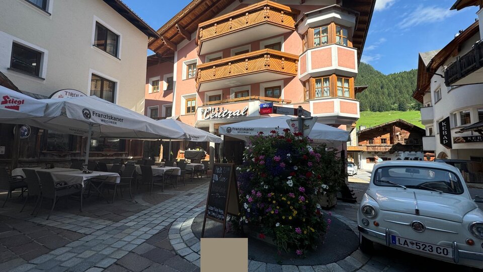 A picturesque town street with charming buildings and a small square. 

In the square, there are flowers and a classic car parked nearby.