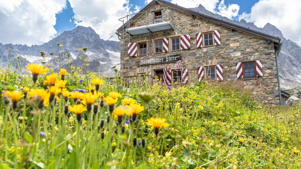 A picturesque stone mountain hut with colorful shutters. The garden is surrounded by yellow flowers and an impressive mountain backdrop.